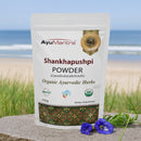 AyuMantra Shankhapushpi Powder packaging with a beach background