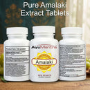 Three white supplement bottles labeled AyuMantra Amalaki, shown on a wooden surface with a blurred natural background. Large text at the top reads 'Pure Amalaki Extract Tablets'.