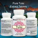 Three white supplement bottles labeled AyuMantra Pure Tulsi Extract Tablets are shown against a scenic mountain-and-river background. The center bottle is front-facing with the brand name and tulsi label visible, while the side bottles show the back panels with ingredients and directions.