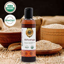 Bottle of AyuMandira Abhyanga Organic Ayurvedic Oil with USDA Organic and OneCert certification logos, surrounded by wooden bowls and a spoon on a dark background.