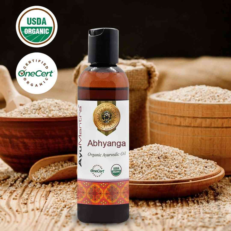 Bottle of AyuMandira Abhyanga Organic Ayurvedic Oil with USDA Organic and OneCert certification logos, surrounded by wooden bowls and a spoon on a dark background.