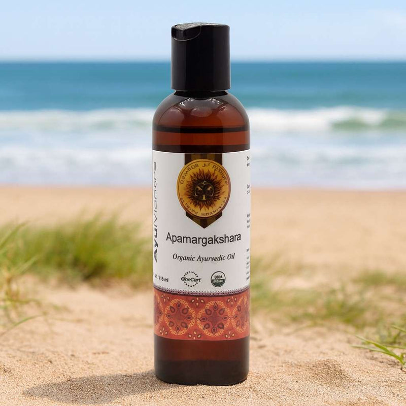 Bottle of Apamargakshara Ayurvedic oil on a sandy beach with ocean in the background