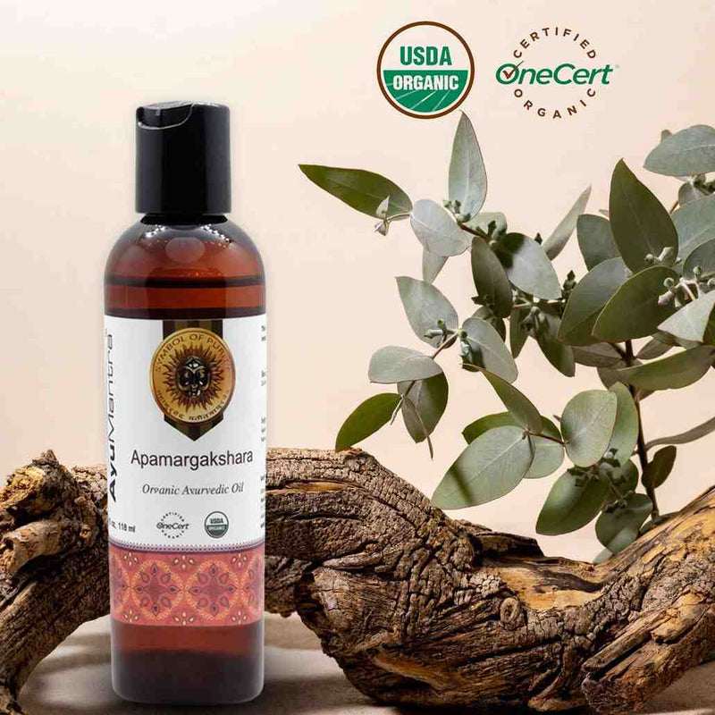 Bottle of Apamargakshara Ayurvedic Oil with USDA Organic and OneCert certification logos, surrounded by eucalyptus leaves and wooden elements.