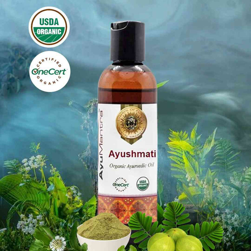 Bottle of Ayushmati organic ayurvedic oil with USDA and OneCert organic certification logos, surrounded by green leaves and plants.