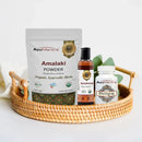 Ayurvedic products including Amalaki powder, oils, and capsules on a woven tray.