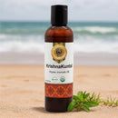 Ayumantra Krishna Kuntal Oil bottle on sandy beach with ocean backdrop and green herbal sprig