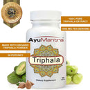 AyuMantra Triphala tablets bottle with benefit highlights and fresh Triphala fruits at the base.
