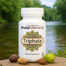 AyuMantra Triphala tablets bottle by a riverside with amla and other herbs in front.