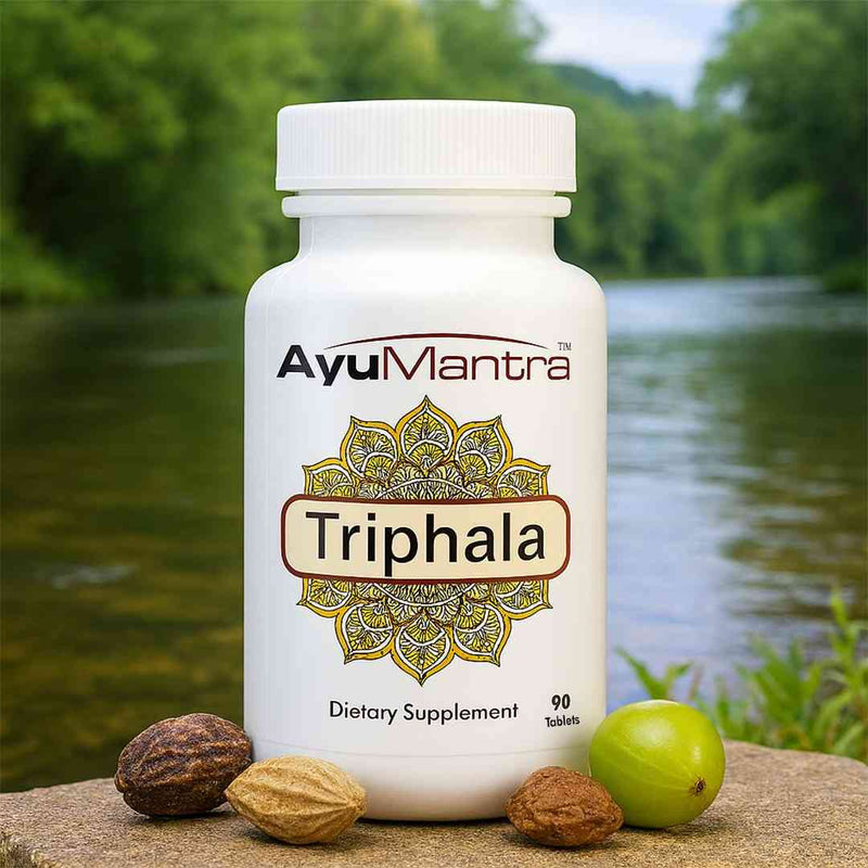 AyuMantra Triphala tablets bottle by a riverside with amla and other herbs in front.